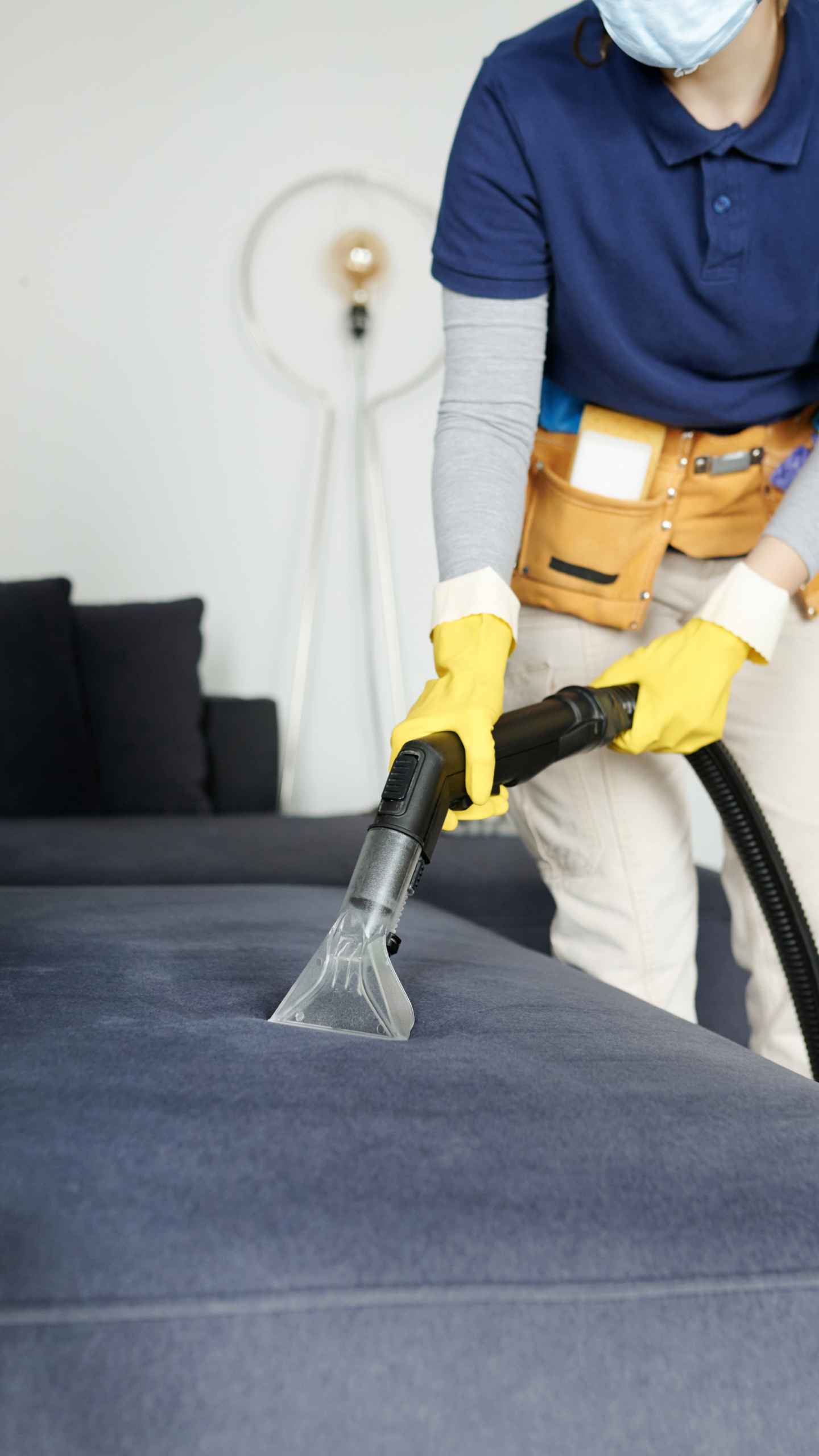 A person wearing a blue shirt, yellow gloves, and a face mask is using a vacuum cleaner attachment to clean a dark blue couch in a well-lit room.